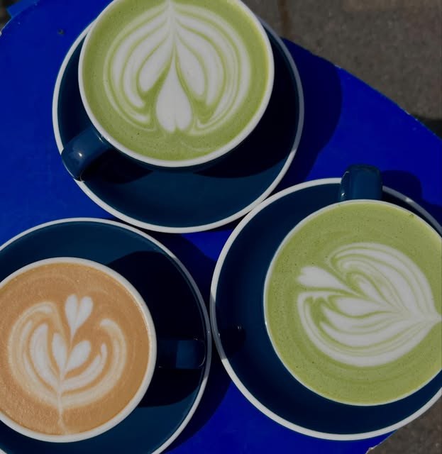 Three lattes with leaf-pattern art, two matcha and one espresso, on a sunlit blue table
